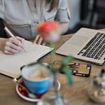Woman at desk writing in a notebook with a computer nearby and a cup of coffee