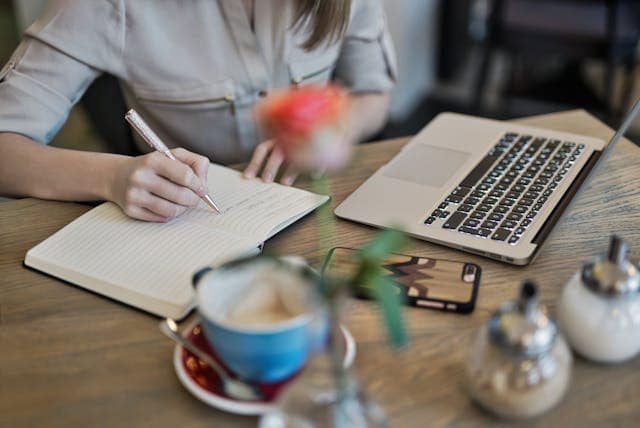 Woman at desk writing in a notebook with a computer nearby and a cup of coffee